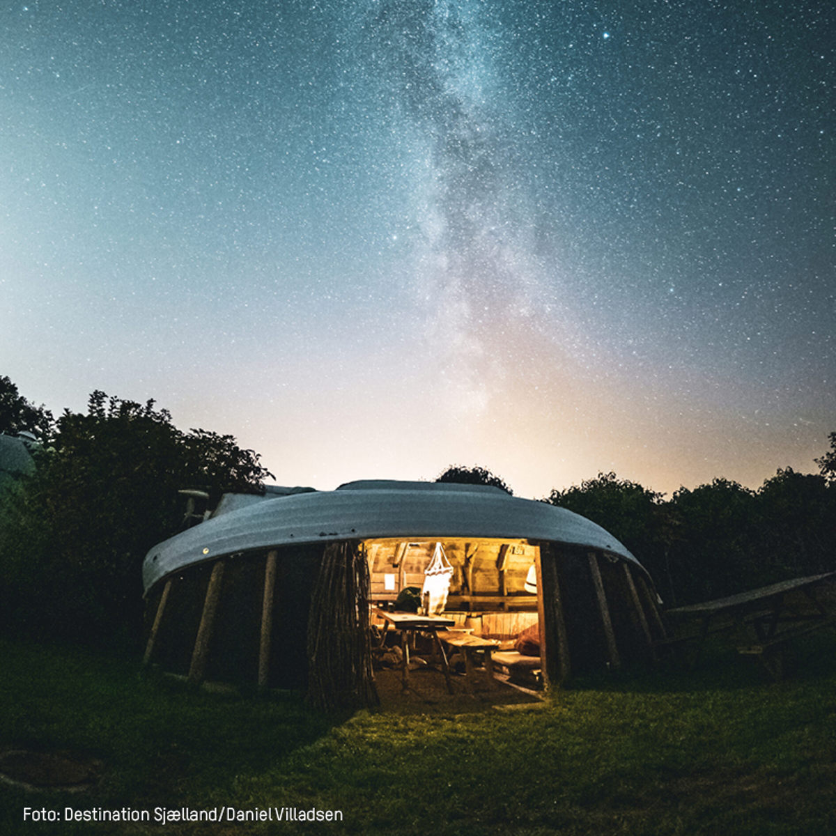 Stjerneklar nat over campinghytte med oplyst interiør, set udefra i en naturskøn have i Danmark.