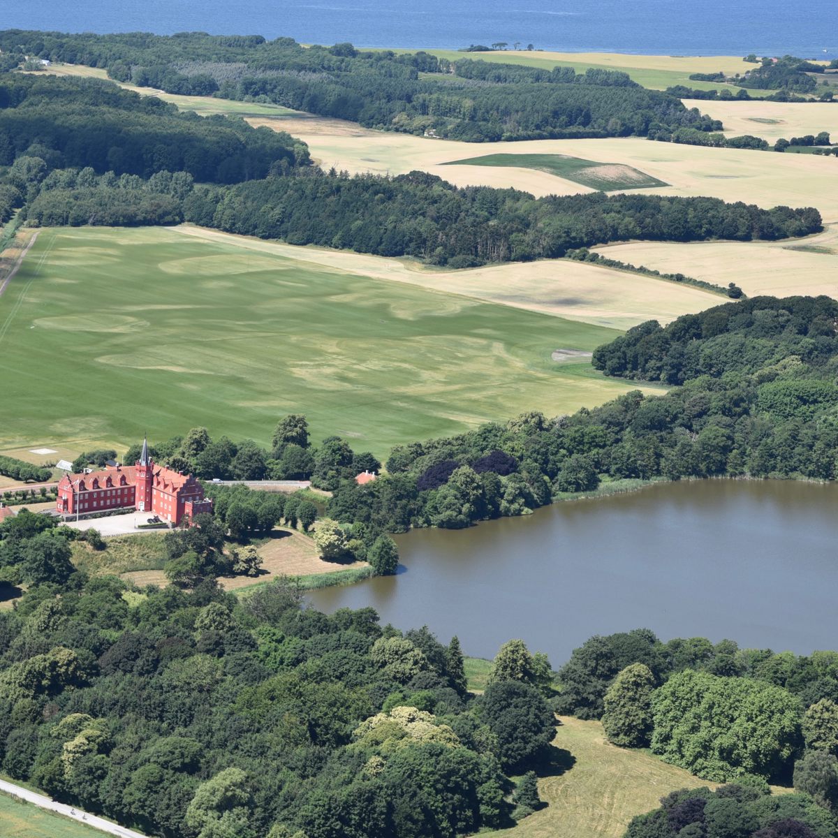 Luftfoto af herregård omgivet af frodige marker og skove nær en sø med udsigt over åbent hav i baggrunden.