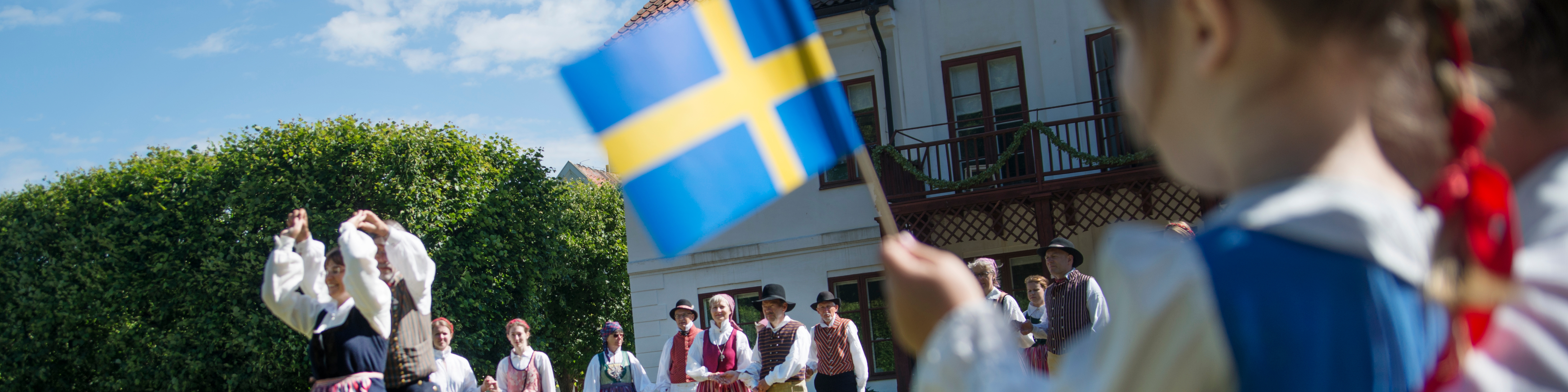 Svensk folkedansgruppe optræder i traditionelt tøj, mens et barn holder det svenske flag i forgrunden.