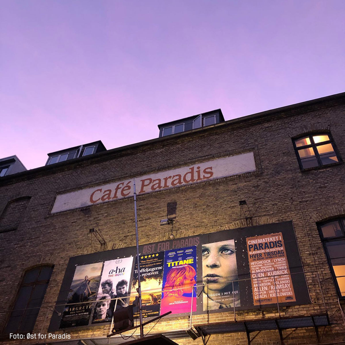 Café Paradis facade med filmplakater under en lilla himmel, set oppefra i skumringen.