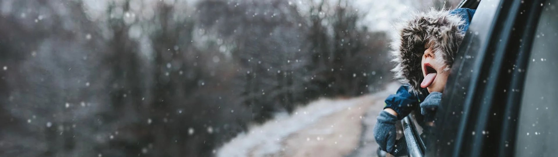 Barn med pelsforet hætte stikker hovedet ud af bilvinduet og fanger snefnug på tungen under snevejr.