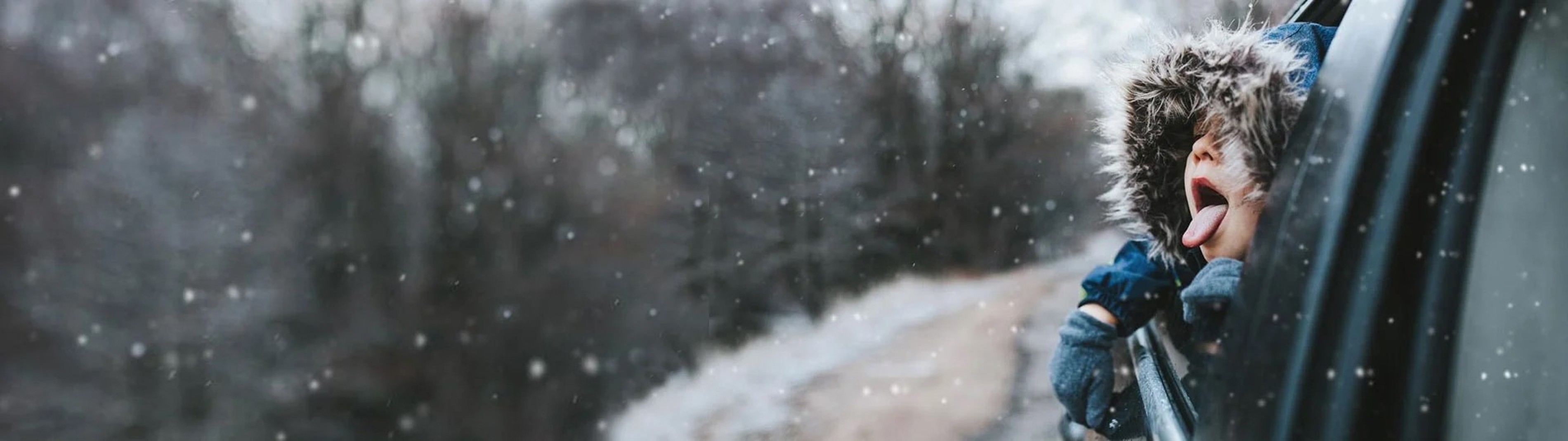 Barn med pelsforet hætte stikker hovedet ud af bilvinduet og fanger snefnug på tungen under snevejr.