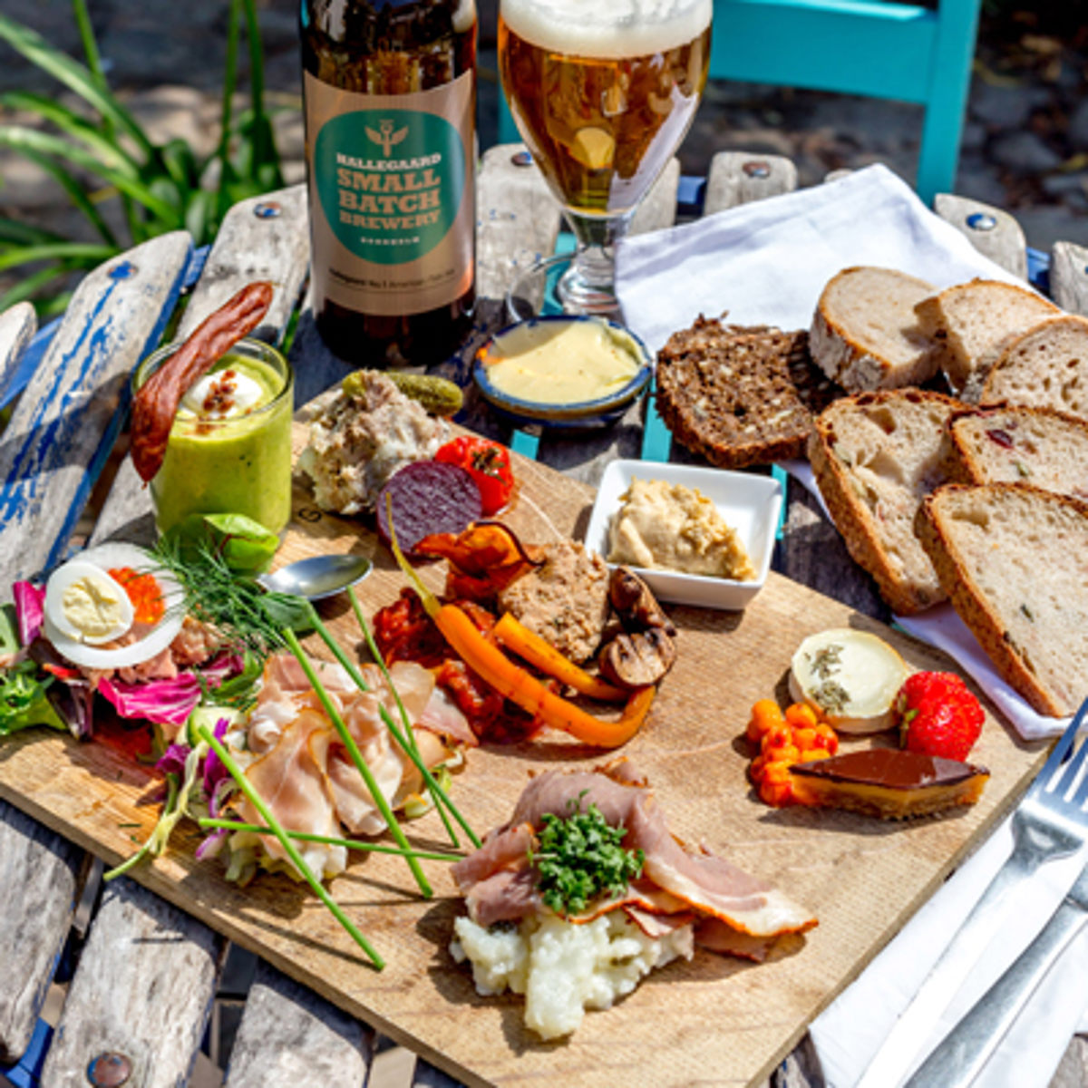 Charcuteribræt med udvalgte oste, skinke, grøntsager og frisk brød serveret udendørs på træbord.