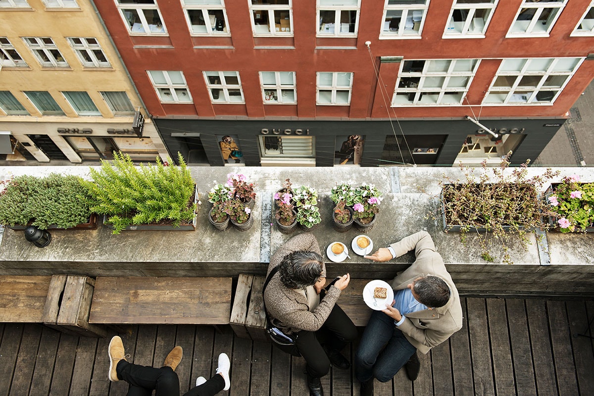 I ett fågelperspektiv ser man ett par som äter och dricker på toppen av en byggnad med utsikt över gatan.