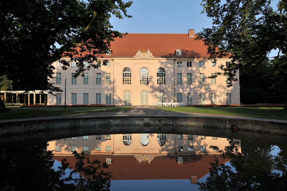 Berlin Schönhausen sett från framsidan med spegelblankt vatten och kvällssol.