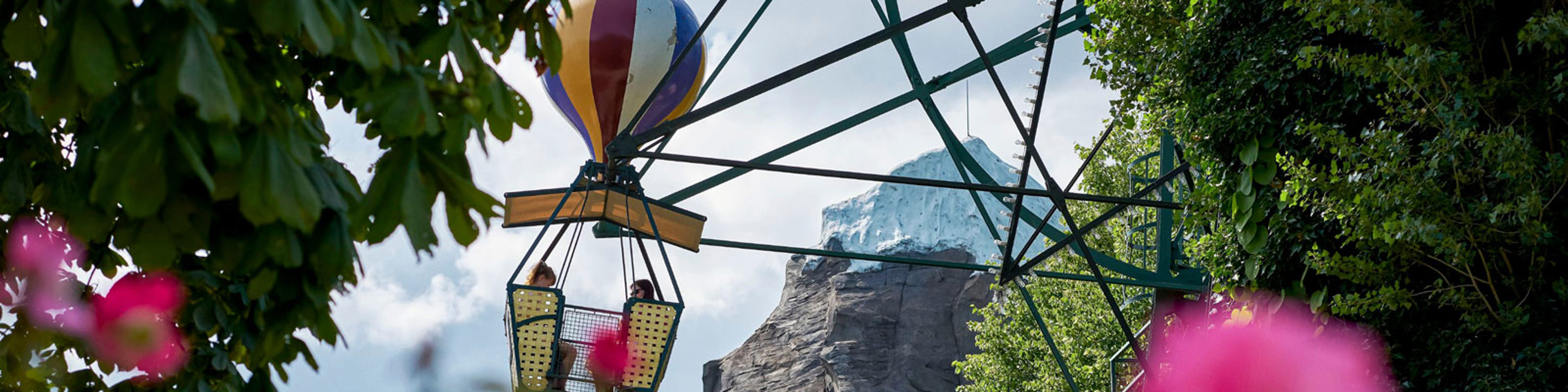 To mennesker i en luftballongynger højt oppe, omgivet af træer og et bjerg i baggrunden i en forlystelsespark.