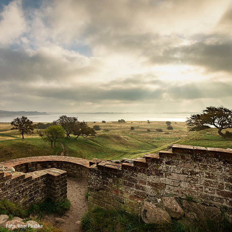 Gammel murstensvæg med udsigt over grønt landskab og hav ved horisonten under dramatisk skyfuld himmel.