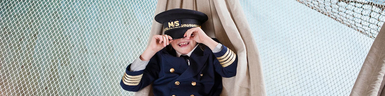 Pojke i sjömanskostym med kaptensmössa leker på Museet For Sjöfart, ler under nätet ombord på en båt.