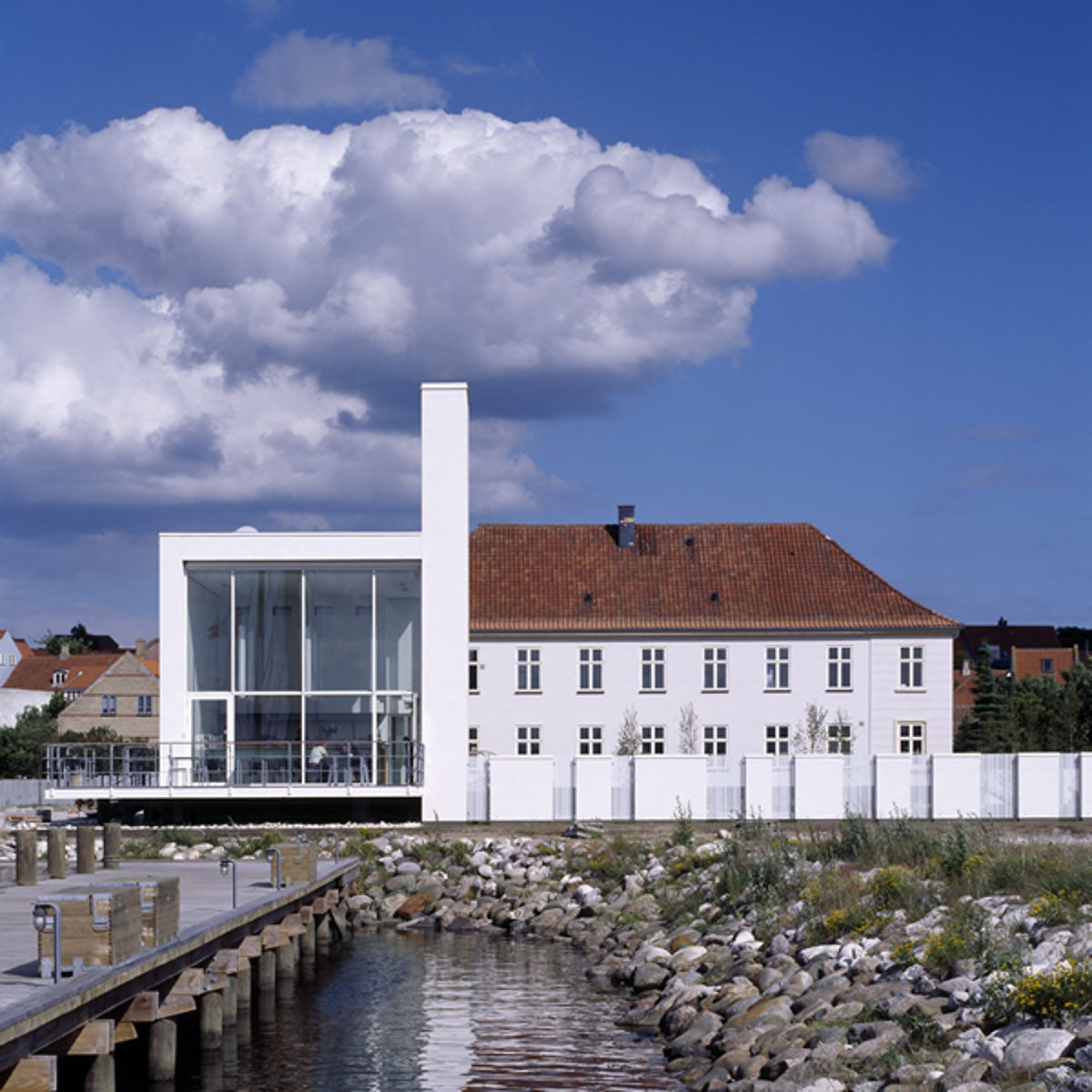 Moderne hvid bygning med glasfacade ved vandkant, skarpt adskilt fra den blå himmel med dramatiske skyer.