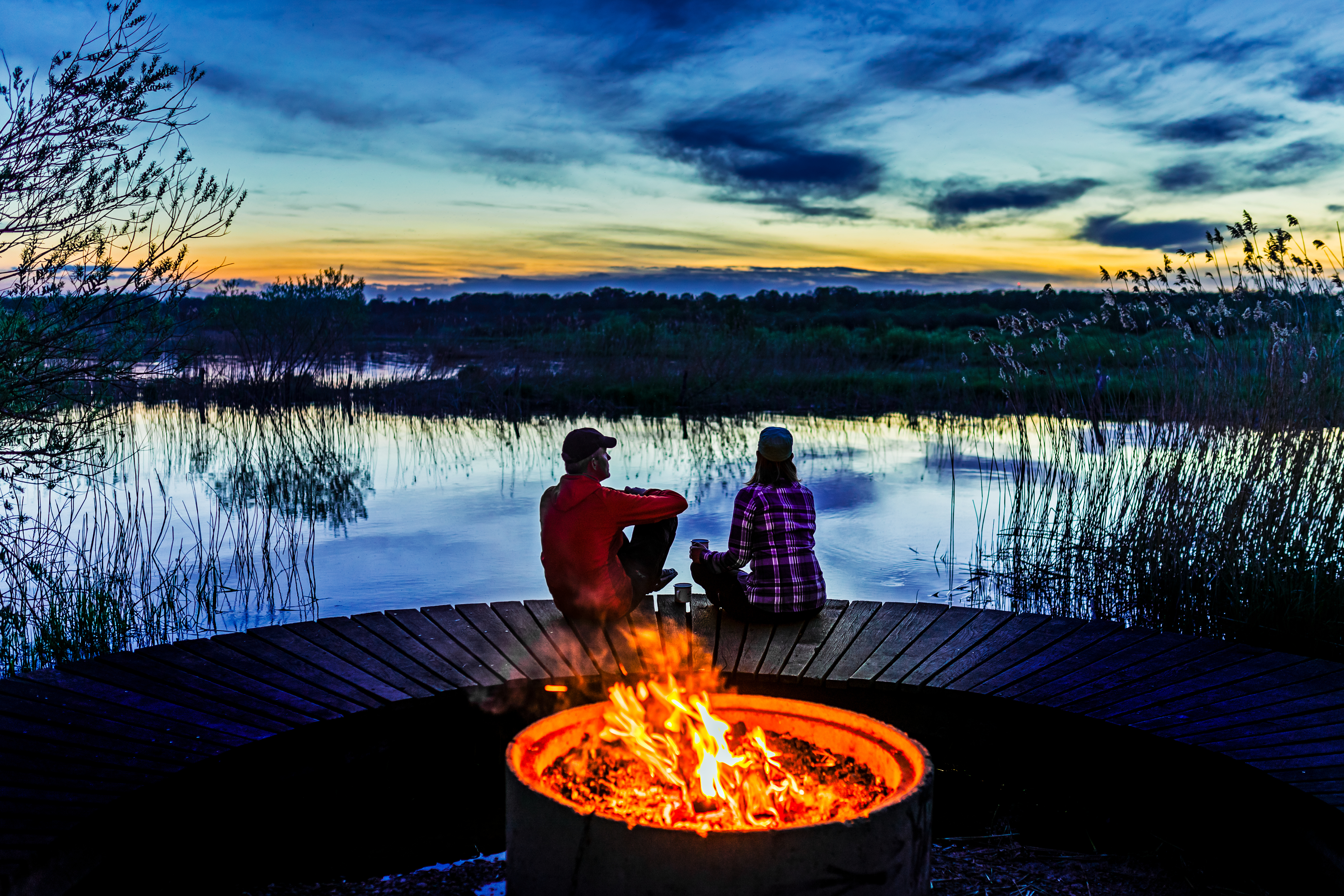 To personer sidder ved et bål på en træbro ved søen i solnedgangens skær.