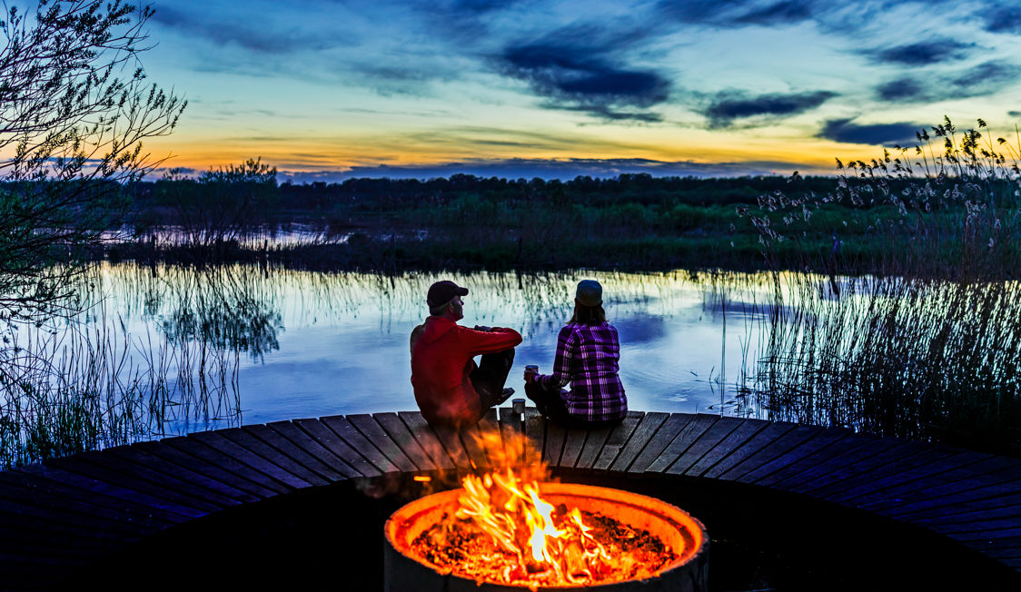 To personer sidder ved et bål på en træbro ved søen i solnedgangens skær.
