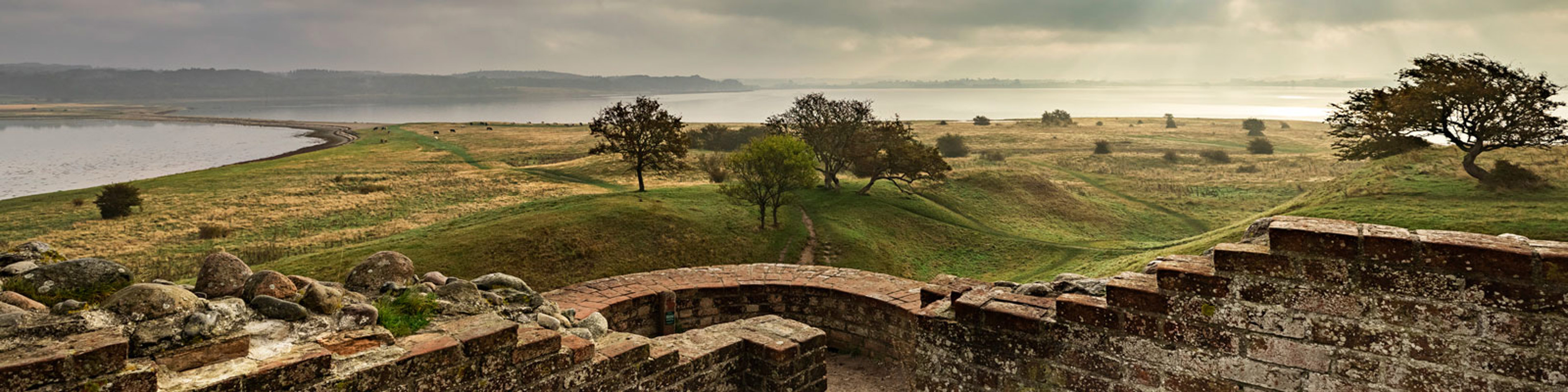 Udsigt over landskab og fjord fra ruinerne af vikingeborgen Trelleborg med skyer i horisonten.