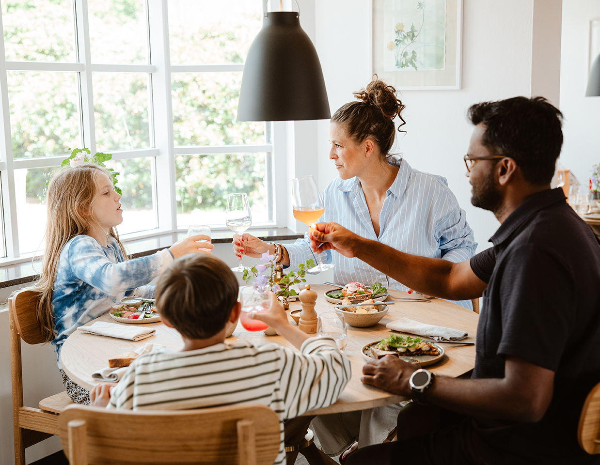 Familie nyder et måltid ved et rundt spisebord, omgivet af lys gennem et stort vindue og moderne indretning.