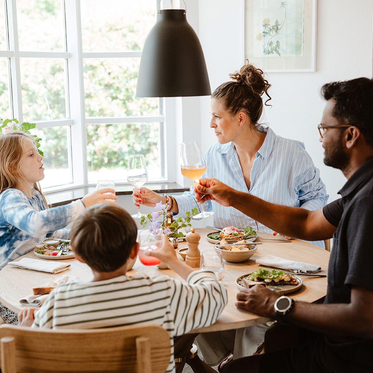 Familie nyder et måltid ved et rundt spisebord, omgivet af lys gennem et stort vindue og moderne indretning.