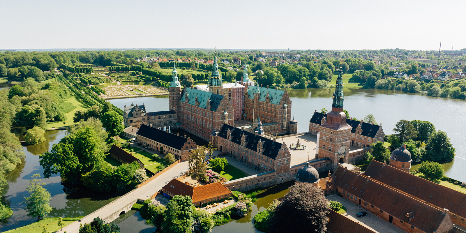 Frederiksborg Slot set fra luften omgivet af frodig natur og spejlblanke søer en solrig sommerdag.