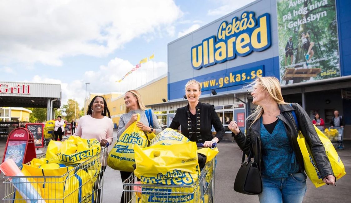 Kunder med indkøbsvogne fyldt med gule poser foran Gekås Ullared-butikken på en solrig dag.