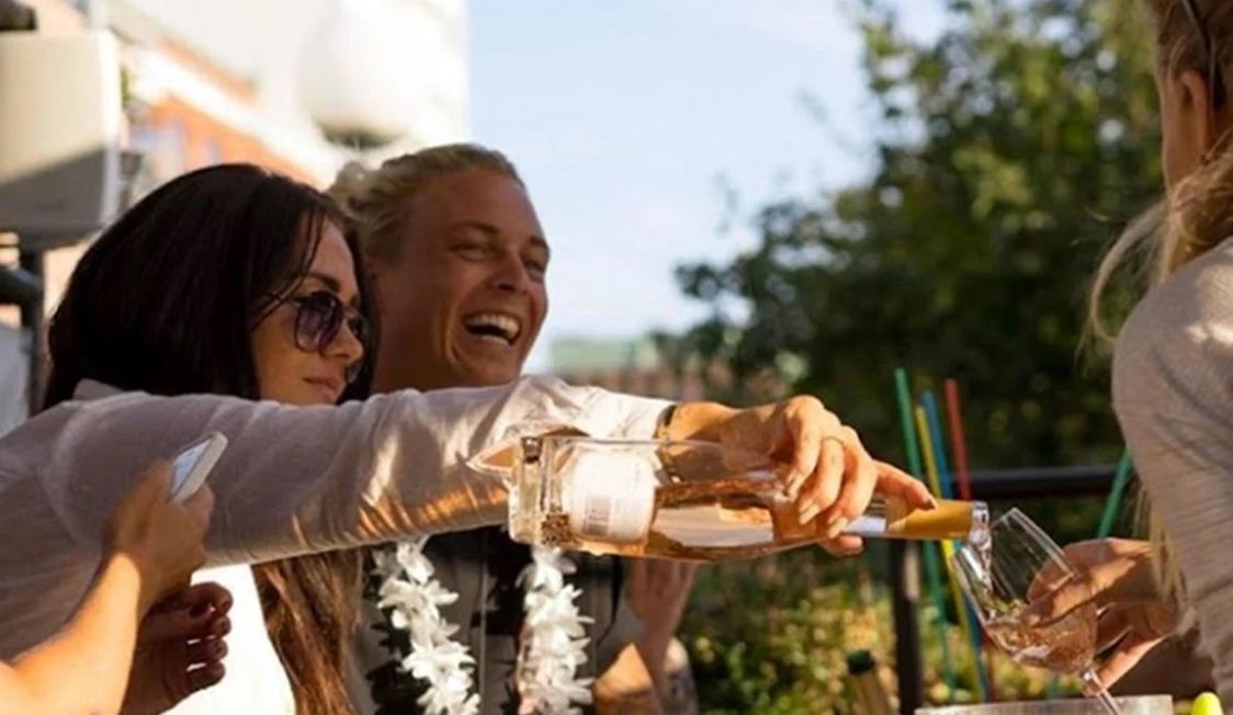 Person hælder rosévin i glas på en solrig terrasse, omgivet af smilende venner, tæt på fritidsaktivitet.