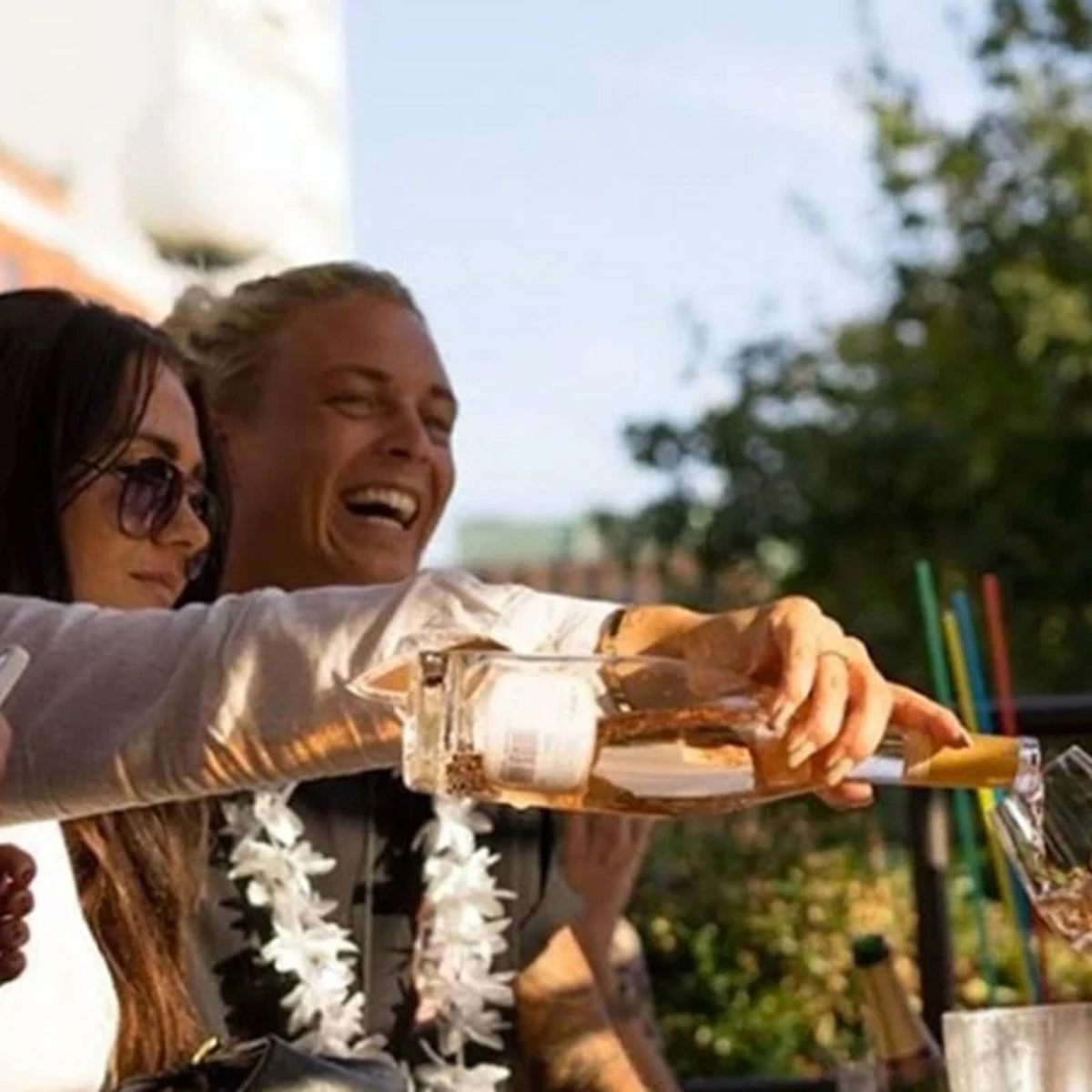 Person hælder rosévin i glas på en solrig terrasse, omgivet af smilende venner, tæt på fritidsaktivitet.