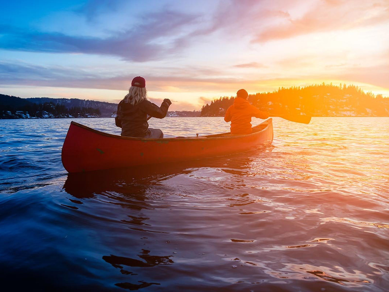 To personer sejler i en rød kano på en sø ved solnedgang, set bagfra i silhuet mod det gyldne lys.