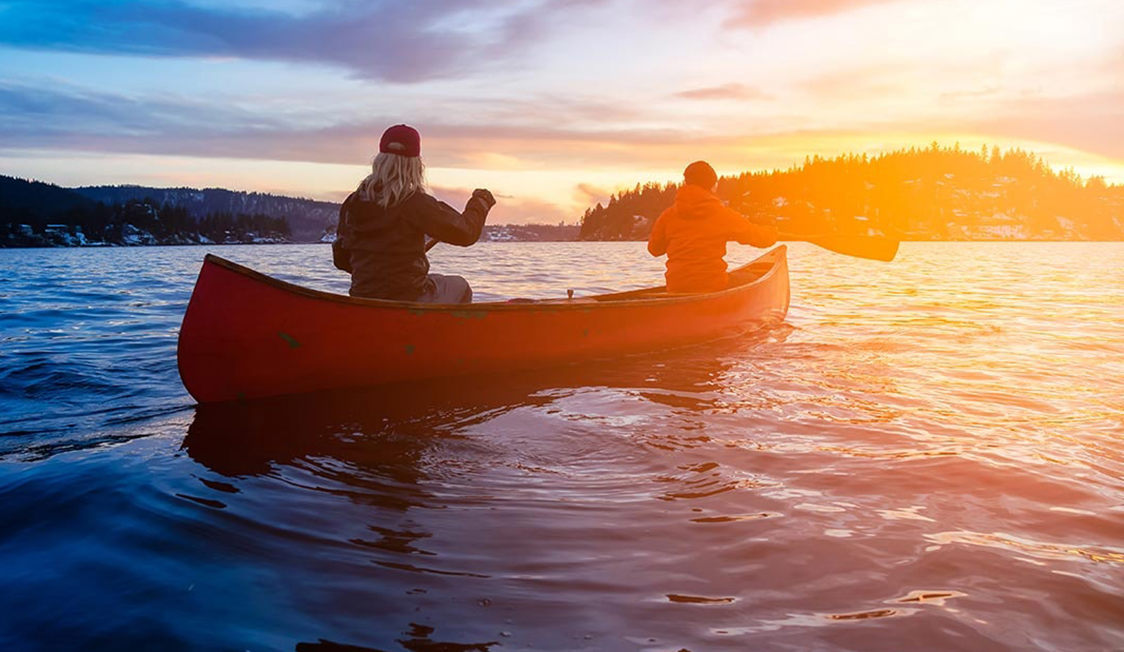 To personer sejler i en rød kano på en sø ved solnedgang, set bagfra i silhuet mod det gyldne lys.