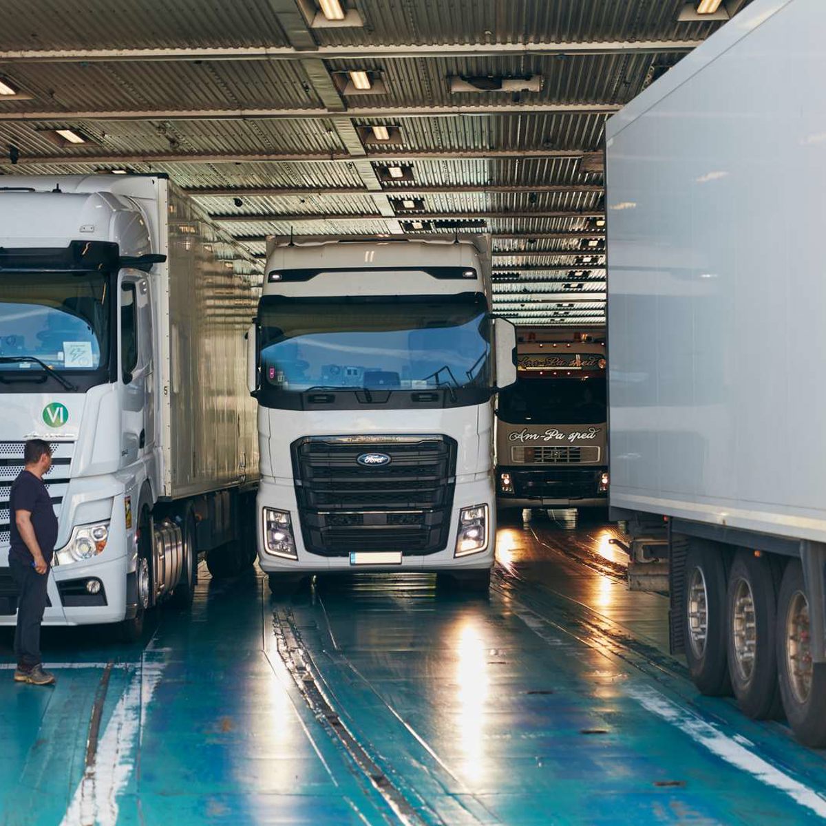 Truck entering ferry cargo hold, surrounded by other trucks, with a man standing beside in illuminated interior.