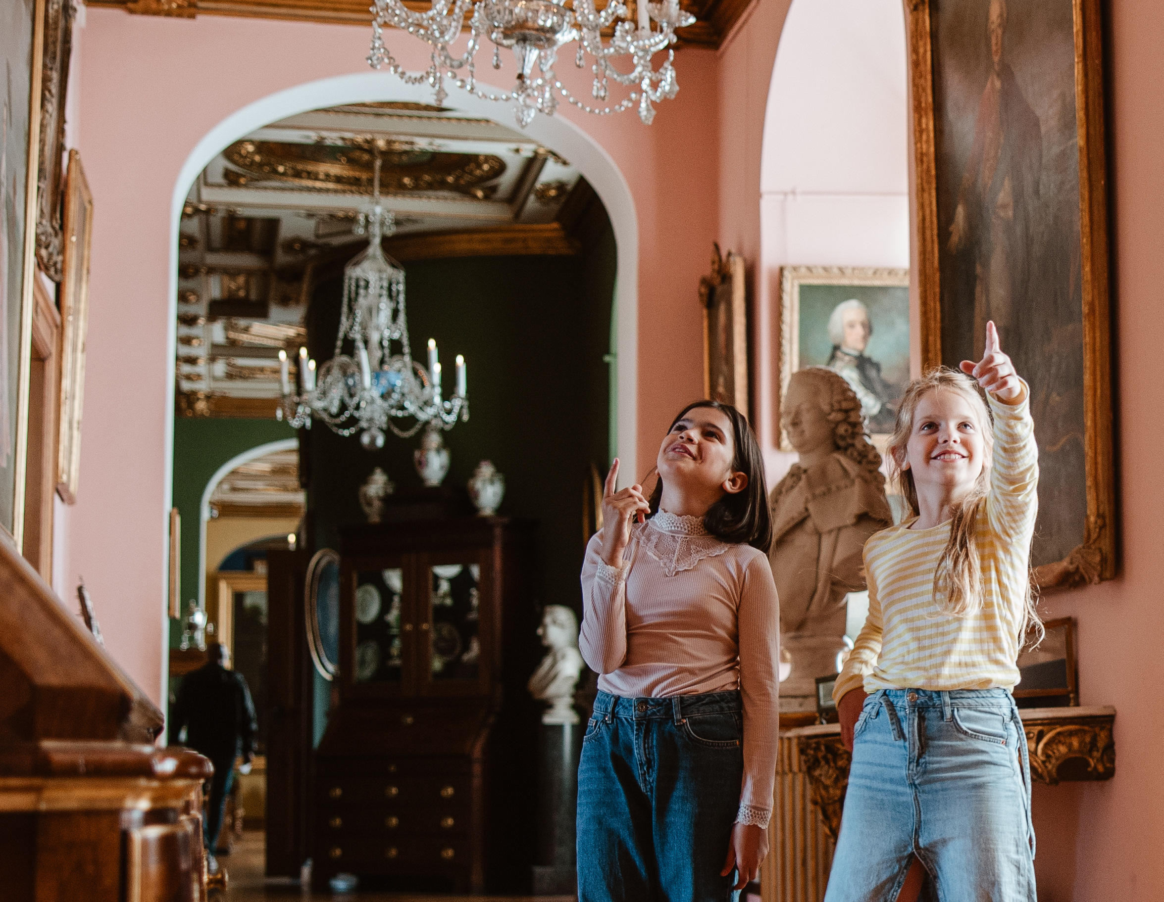 To piger peger opad i en elegant kunstudstilling med lysekrone og buster i baggrunden.