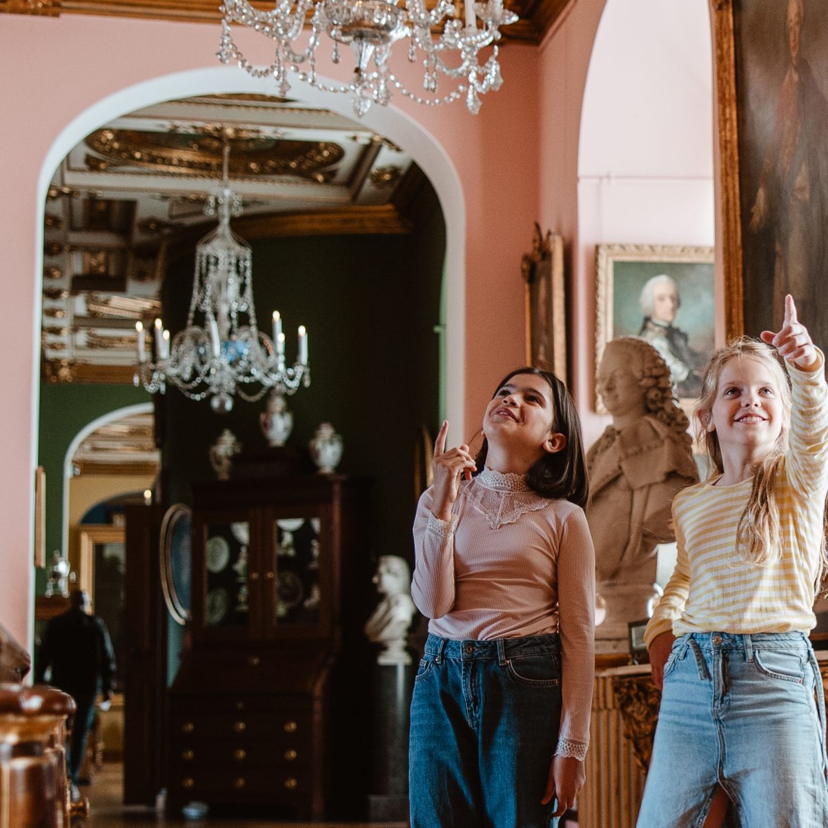 To piger peger opad i en elegant kunstudstilling med lysekrone og buster i baggrunden.