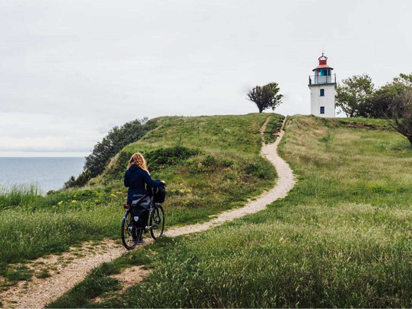 Cyklist på grusväg mot fyr i Hundested med grönskande landskap och hav i fjärran.