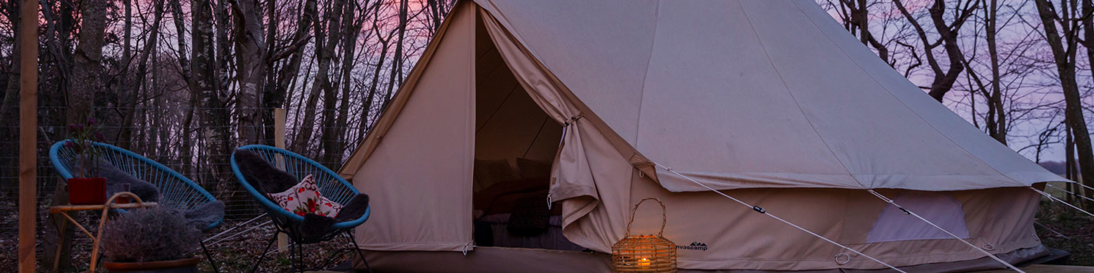 Beige telt opsat i skovmiljø ved solnedgang, indgangslyset skaber en hyggelig atmosfære sammen med naturrattan lampe.