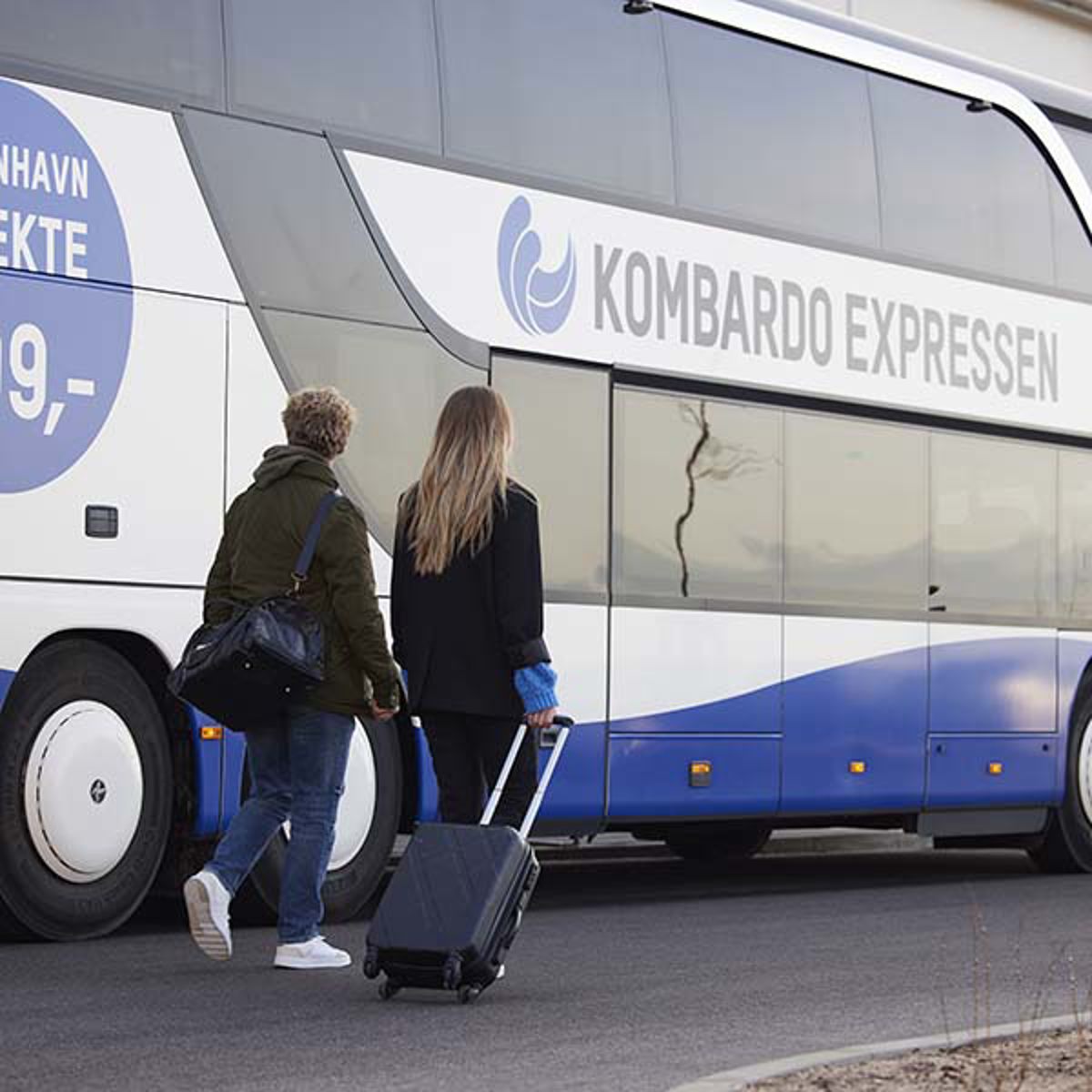 To personer går mod en Kombardo Expressen-bus med kufferter på parkeringsplads.