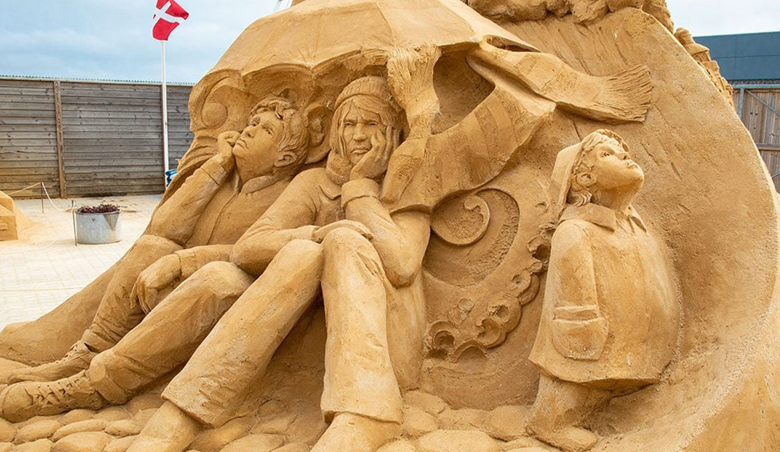 Sandskulpturen Hundested föreställer tre personer under ett paraply, skapad av sand med ett detaljerat hantverk.