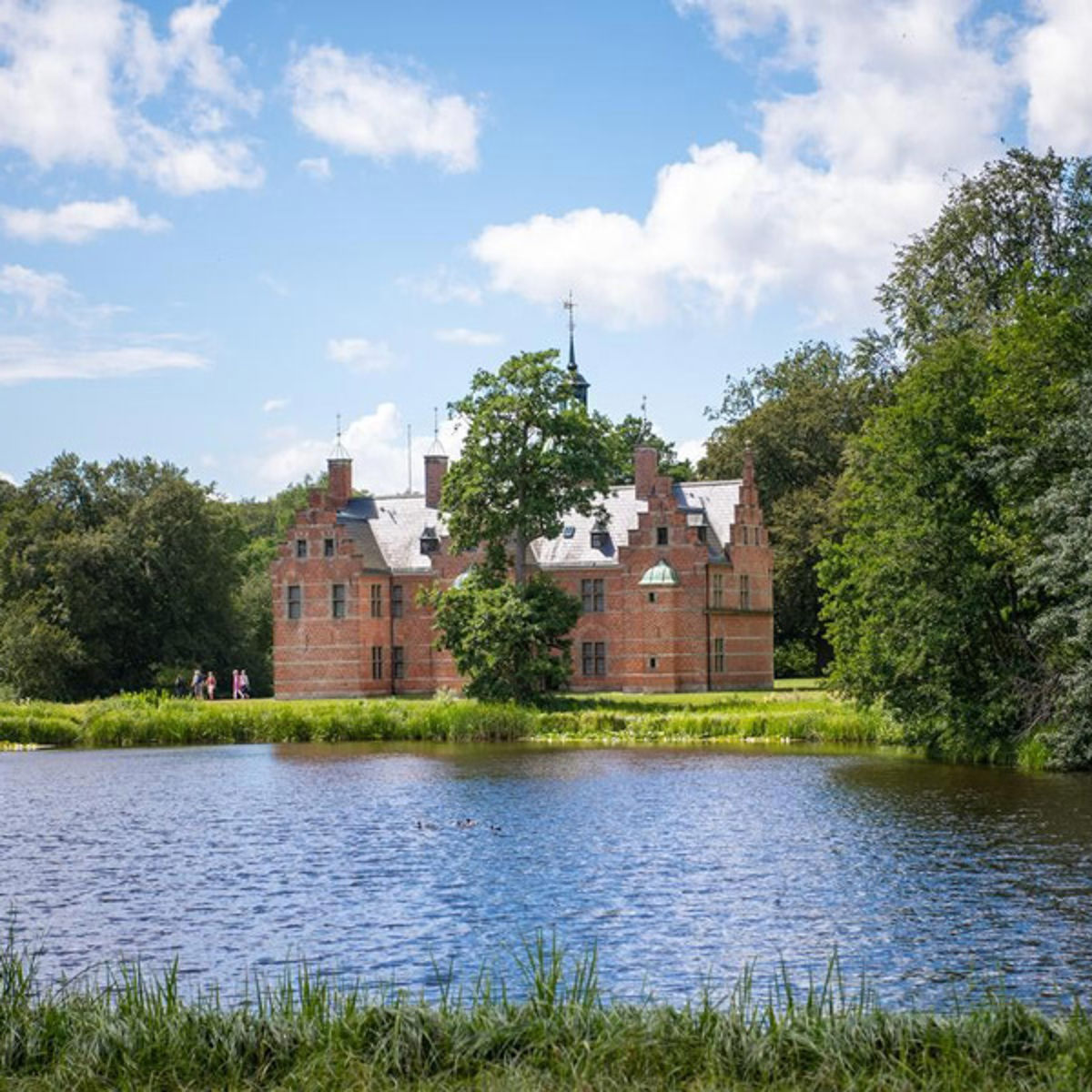 Hilleröds Slott vid sjön en solig dag med moln på himlen och grönska i förgrunden.