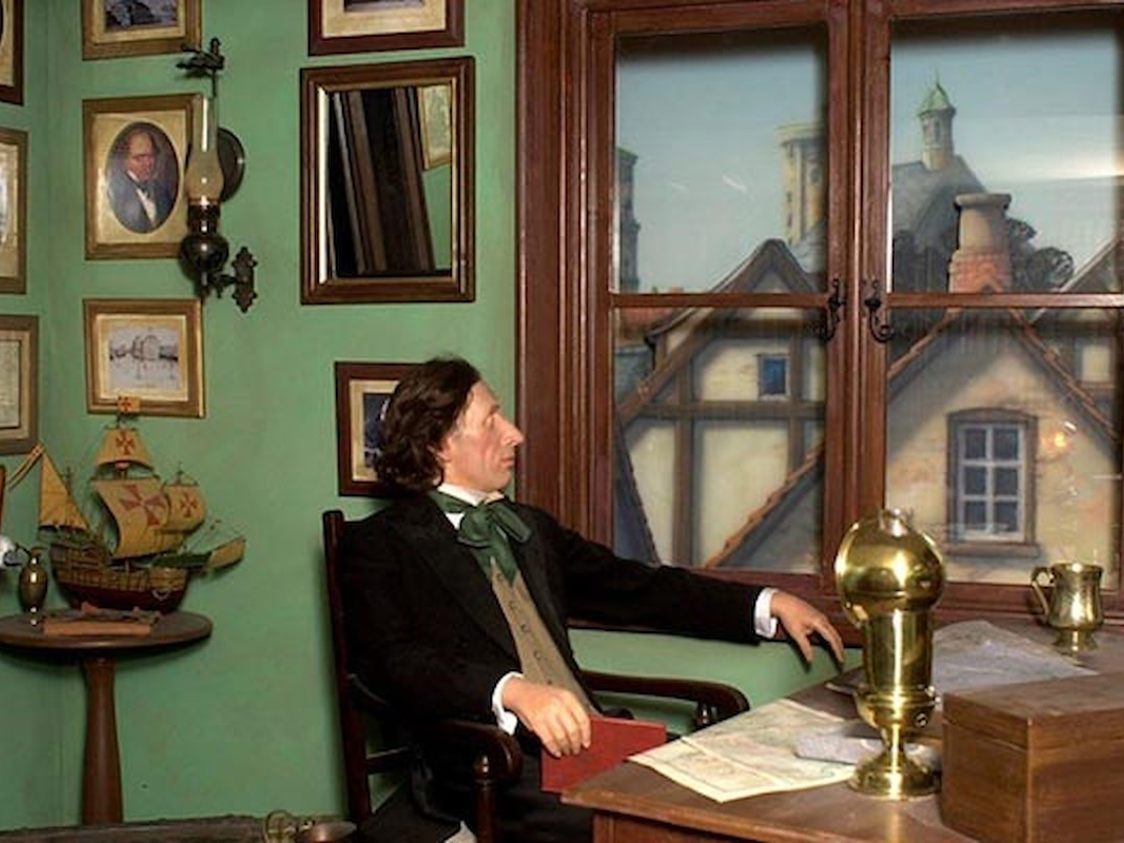 H.C. Andersen sittande vid ett grönt skrivbord, omgiven av tavlor och modeller, blickar ut genom fönstret.