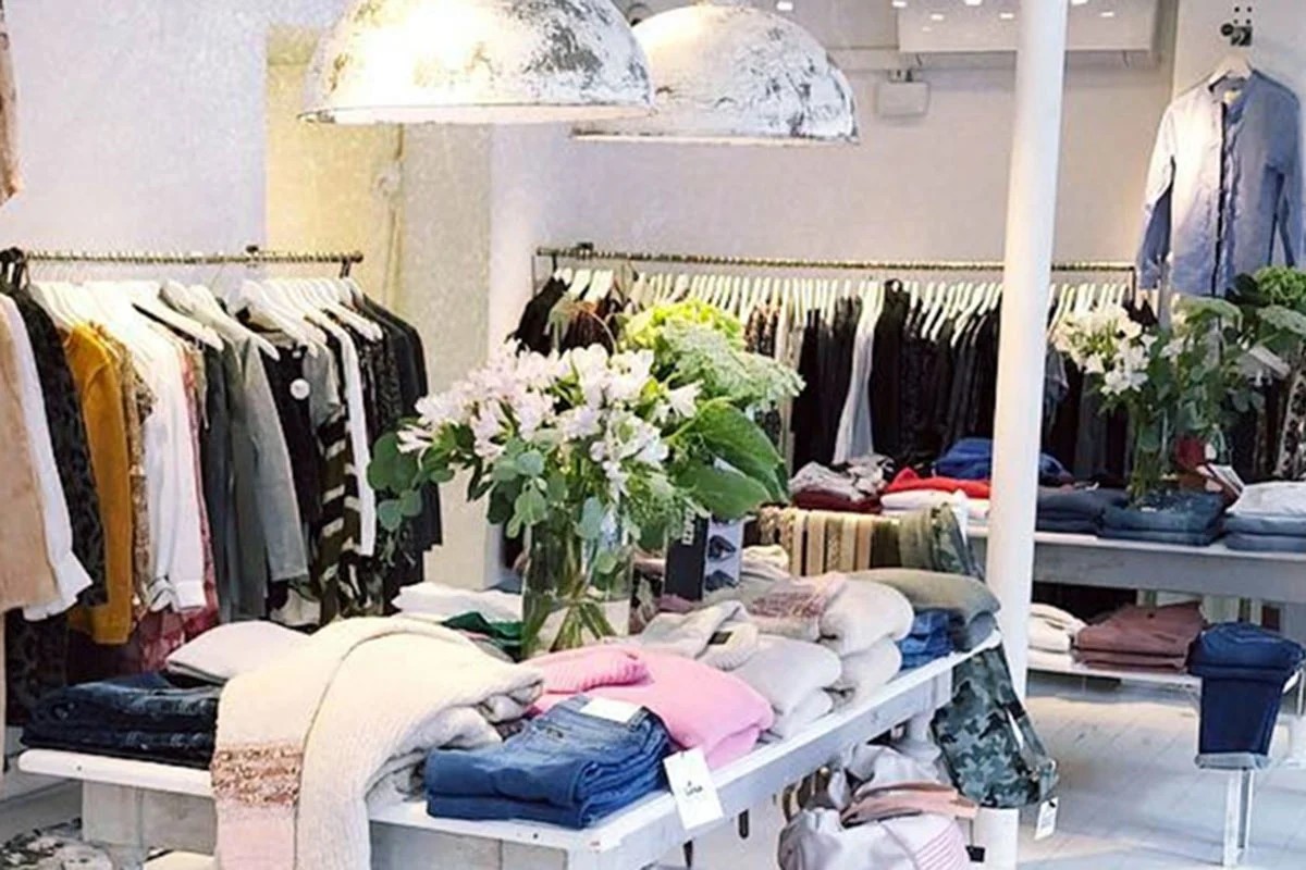 Beklædningsbutiks interiør med tøj på borde og bøjler, lysindfald og blomster i vase i centrum.