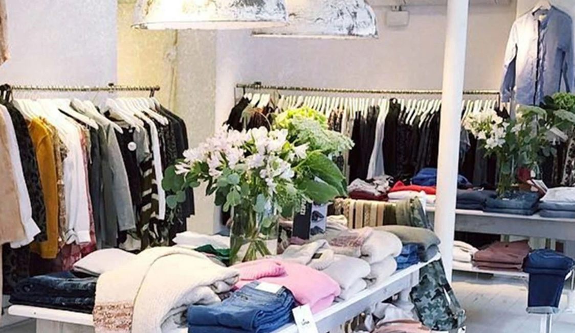 Beklædningsbutiks interiør med tøj på borde og bøjler, lysindfald og blomster i vase i centrum.