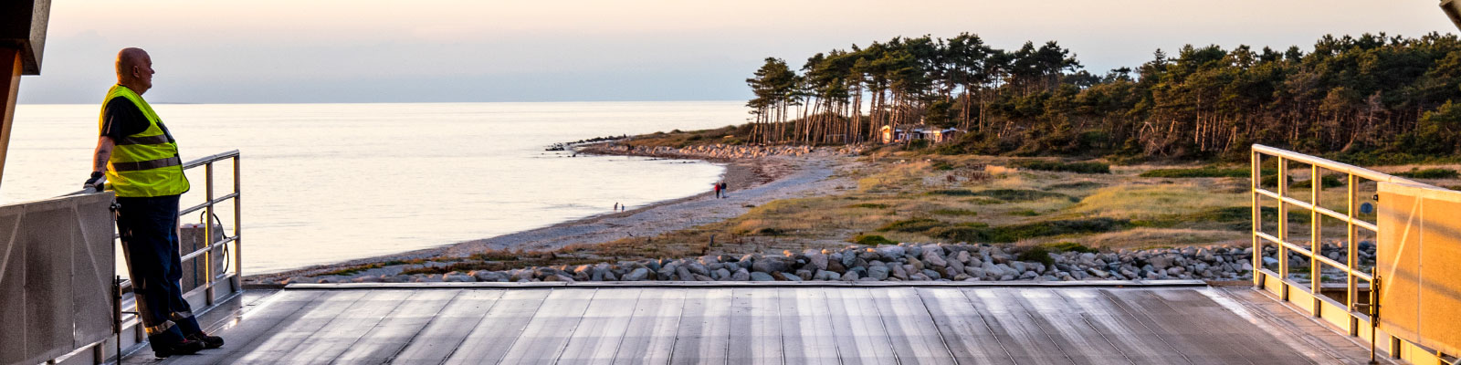 Beklædt arbejder i fluorescerende vest står på færgedæk og ser ud over kystlandskabet ved solnedgang.