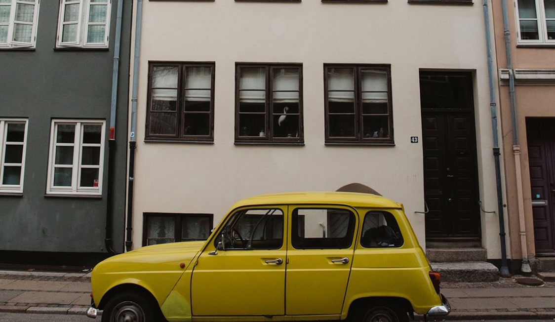 Gul bil parkerad framför ett beige hyreshus i en urban miljö.