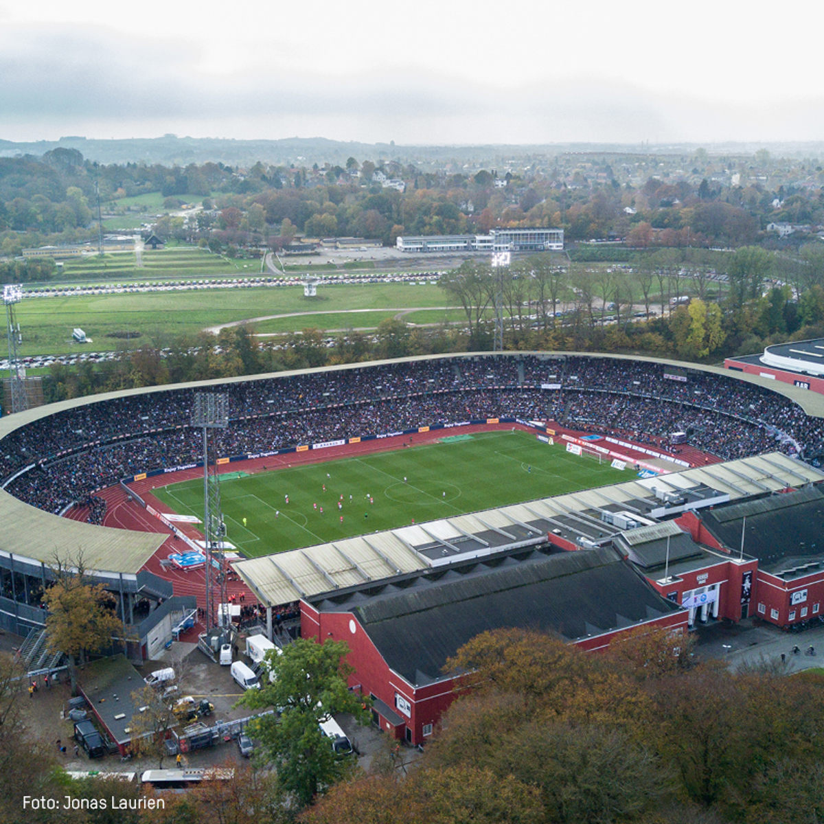 Luftfoto af et fyldt stadion under en fodboldkamp, set fra oven med grønne områder i baggrunden.