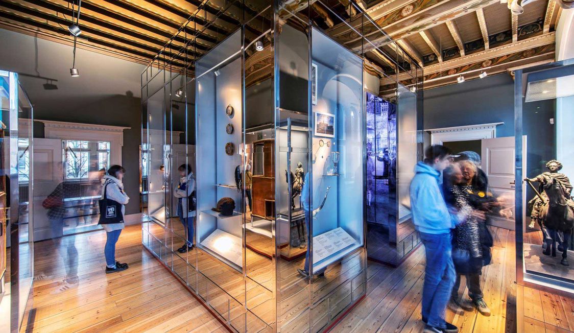 Museumsgæster betragter udstillinger i moderne glasmontre på et historisk trægulv.