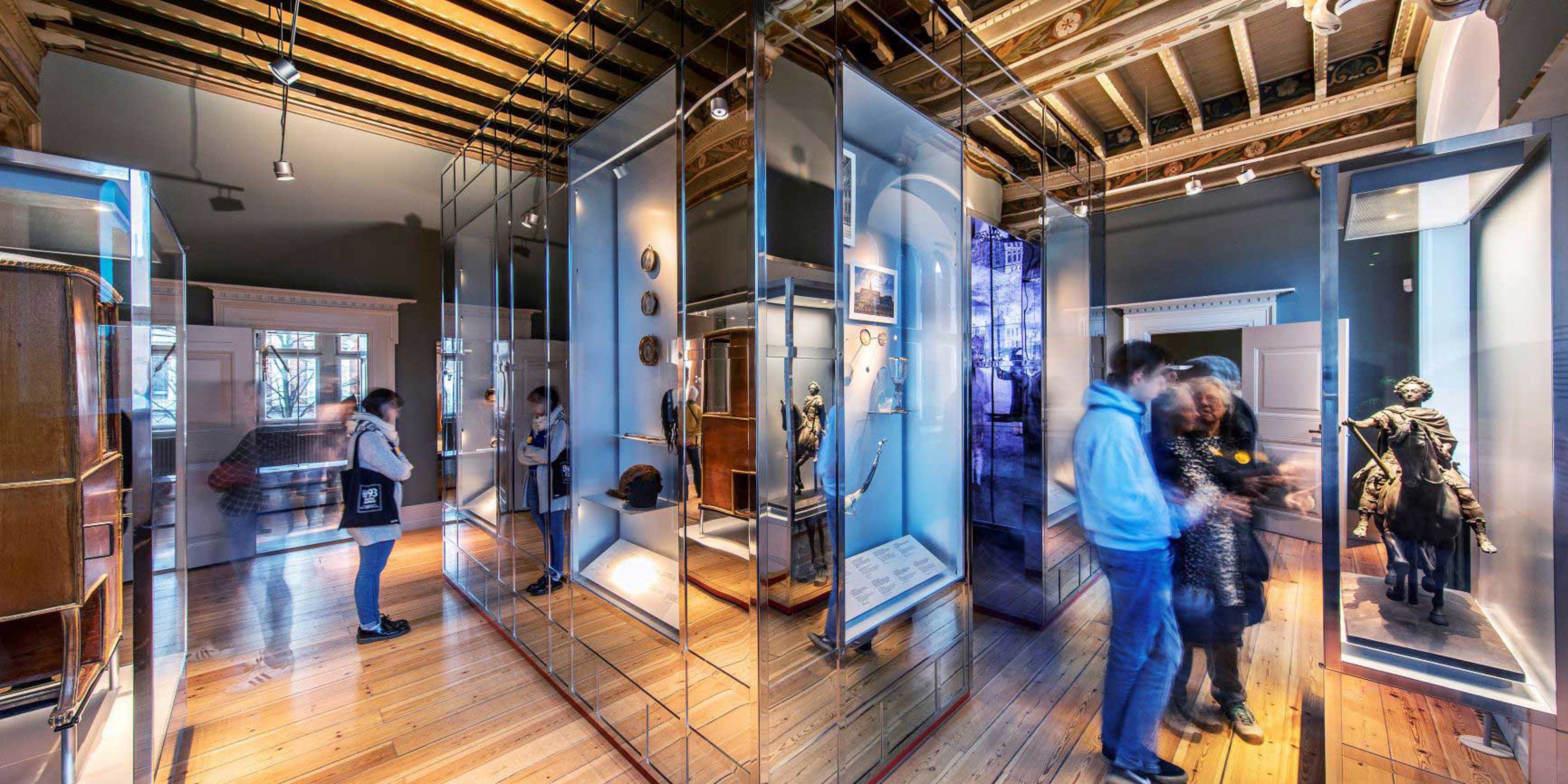 Museumsgæster betragter udstillinger i moderne glasmontre på et historisk trægulv.