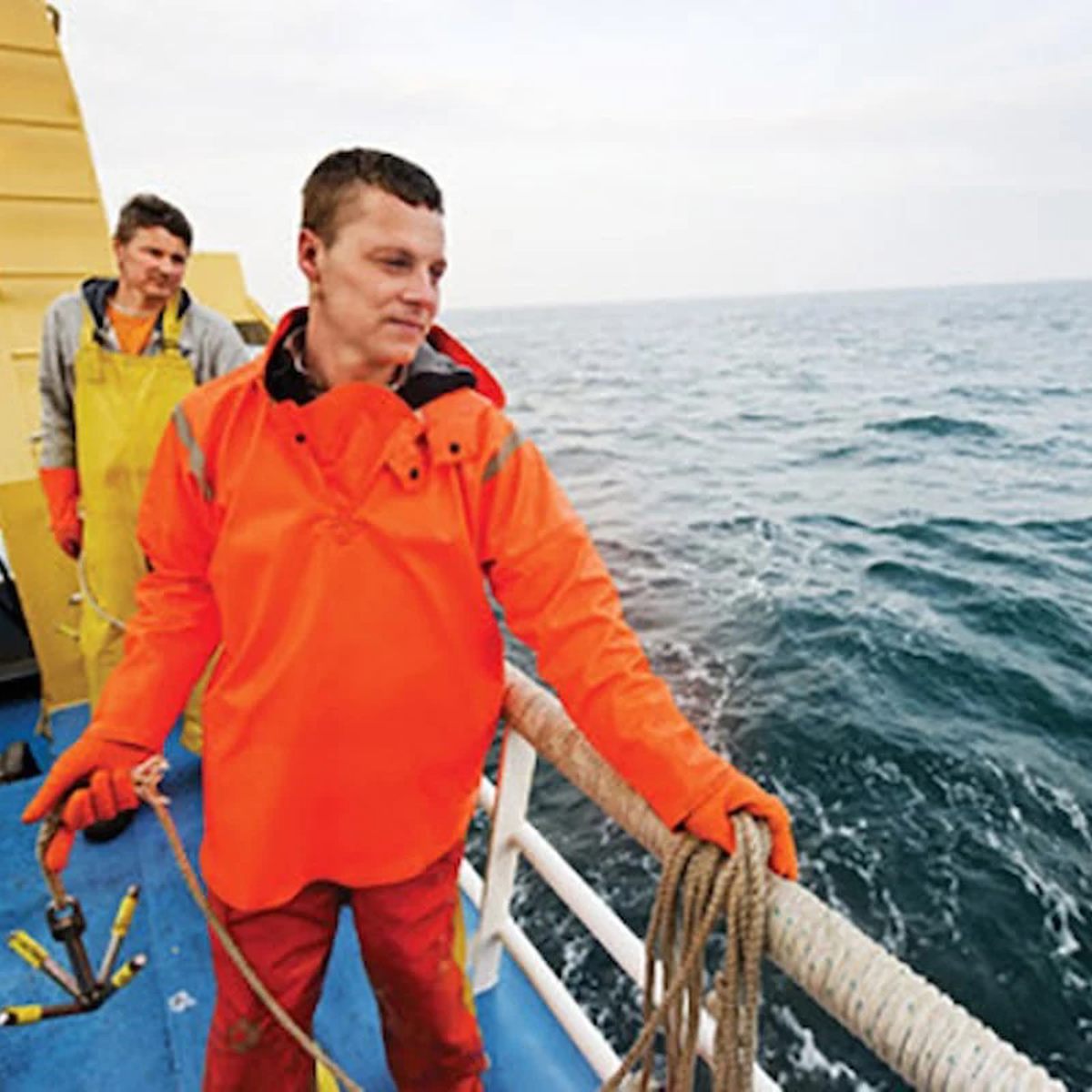 Mand iført orange regnjakke arbejder på fiskerbåd på havet, holder tov ved rælingen i overskyet vejr.