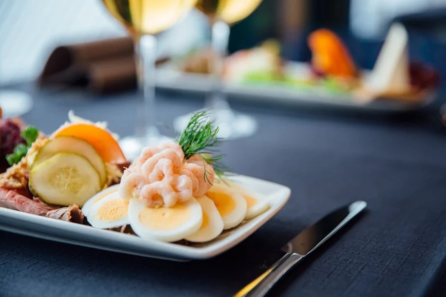 Rejemad med æg, dild og agurkeskiver serveret på hvid tallerken mod en mørk dug på restaurantbord.