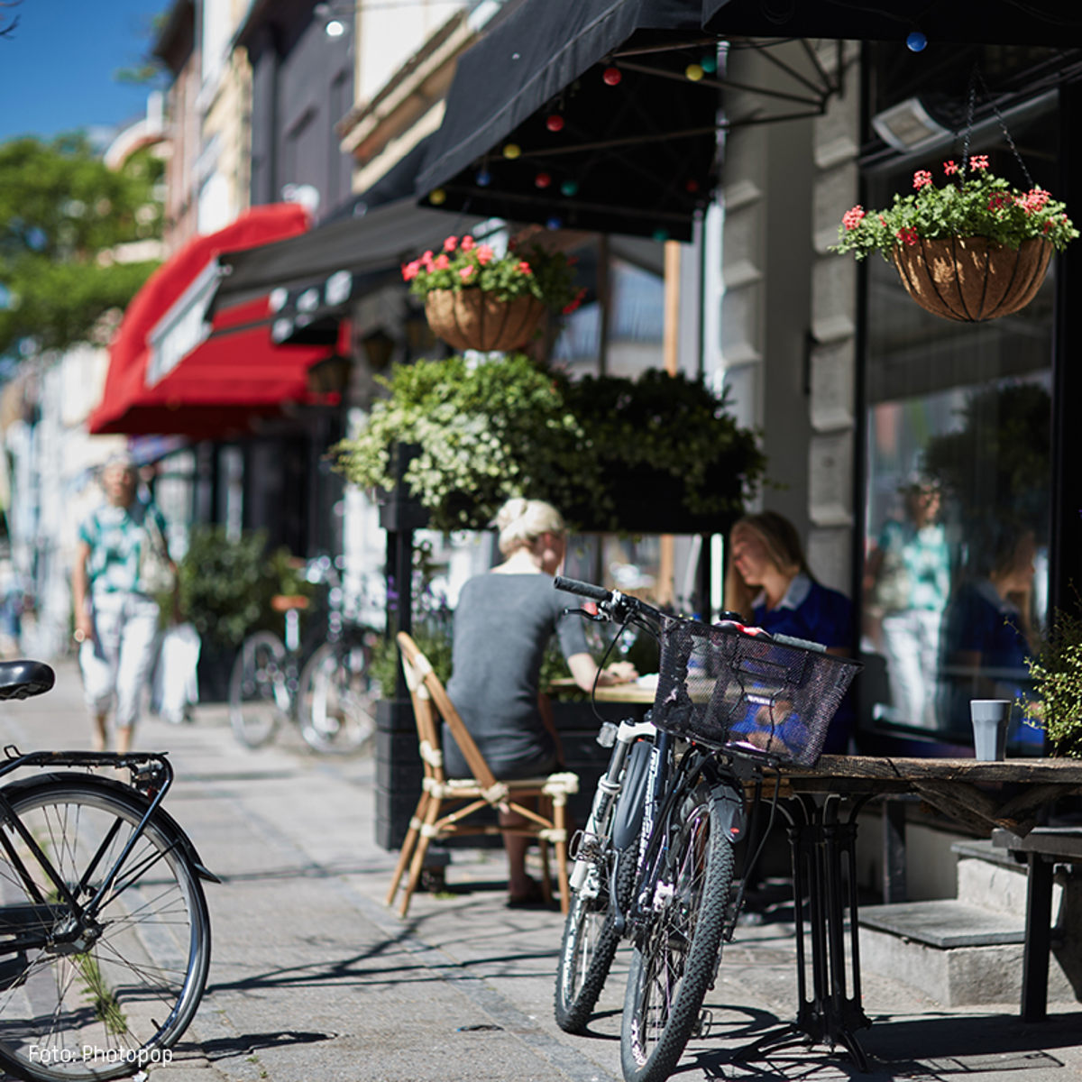 Cykel parkeret ved cafébord på fortovet i en solrig gade, hvor folk sidder og nyder udendørs servering.