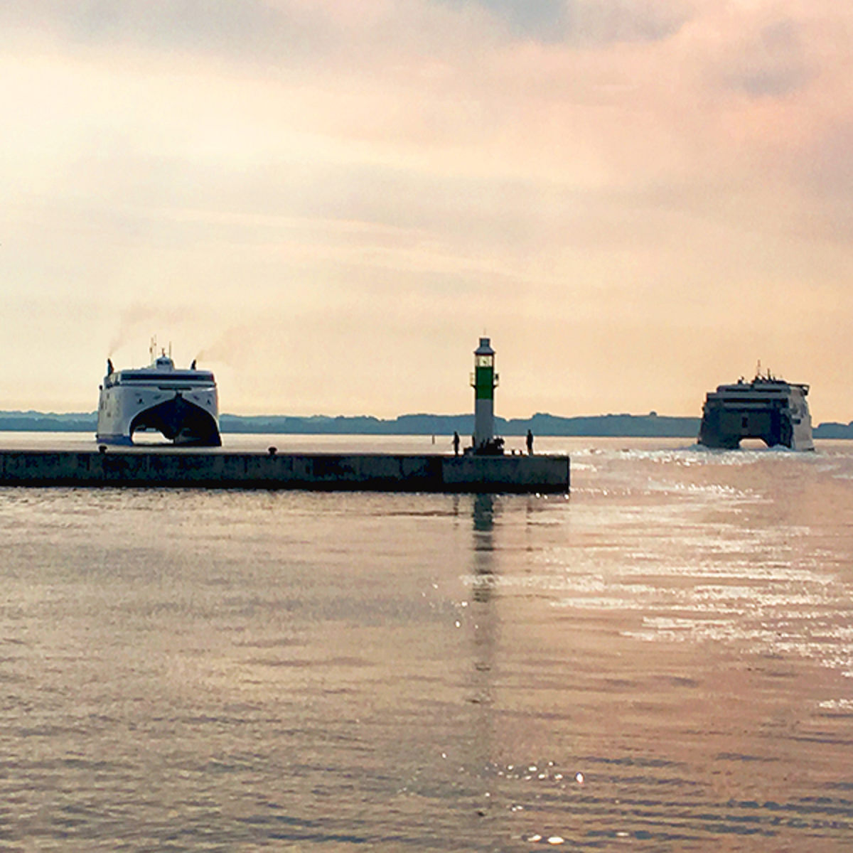 To færger ankommer til havn ved en mole med en grøn fyrtårn i solnedgangen.