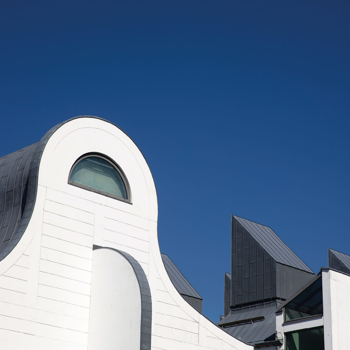 Moderne arkitektur med buet hvid facade og skulpturelle skifertage under en klar blå himmel.