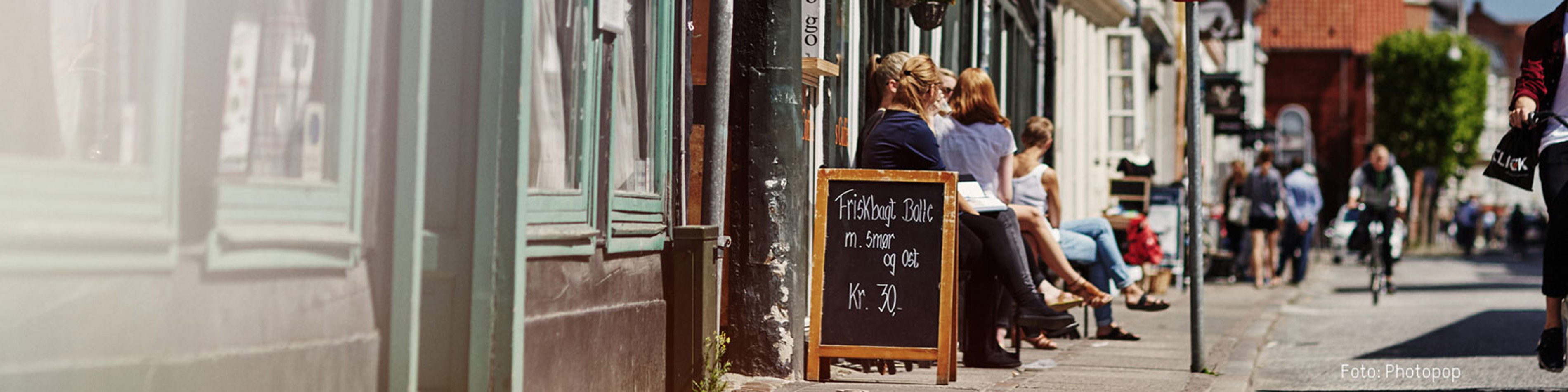 Folk sidder udenfor en café på en solrig dag, med en tavle der annoncerer friskbagte boller med smør og ost.