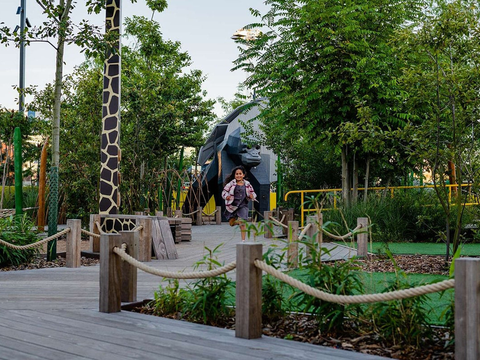 Barn løber glad på en sti i en legeplads med grønne planter og et stort gorillaskulptur i baggrunden.