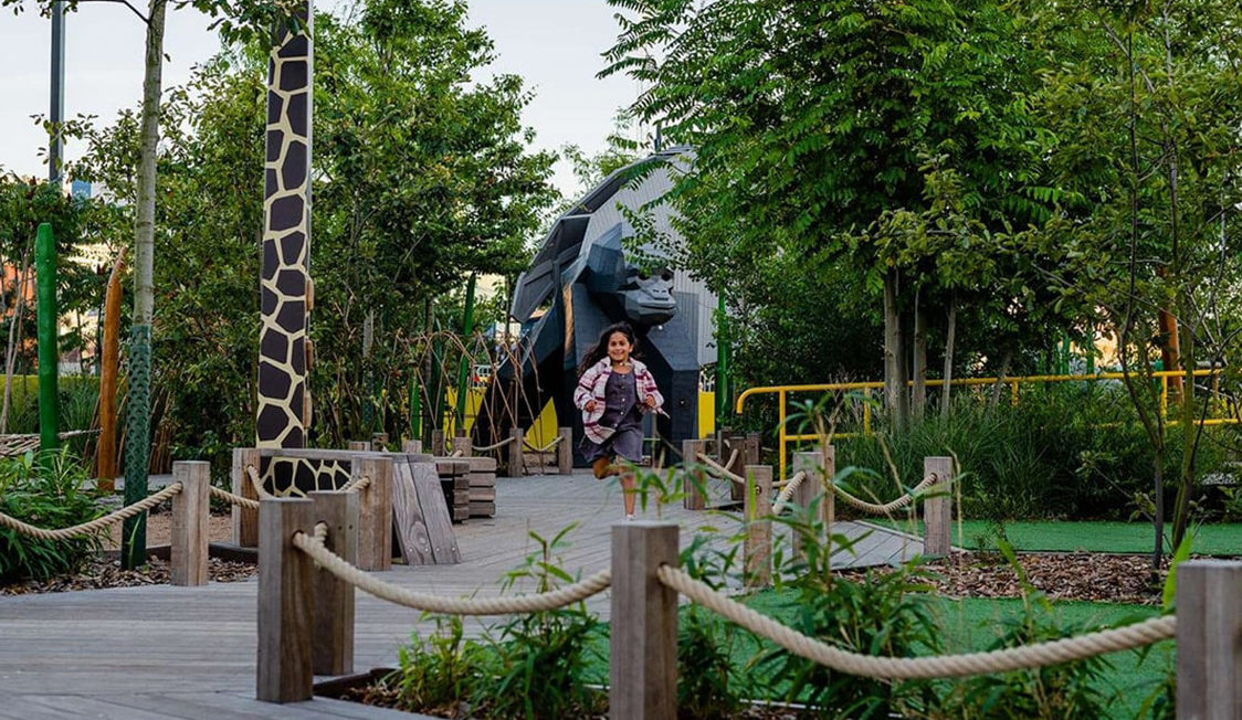 Barn løber glad på en sti i en legeplads med grønne planter og et stort gorillaskulptur i baggrunden.