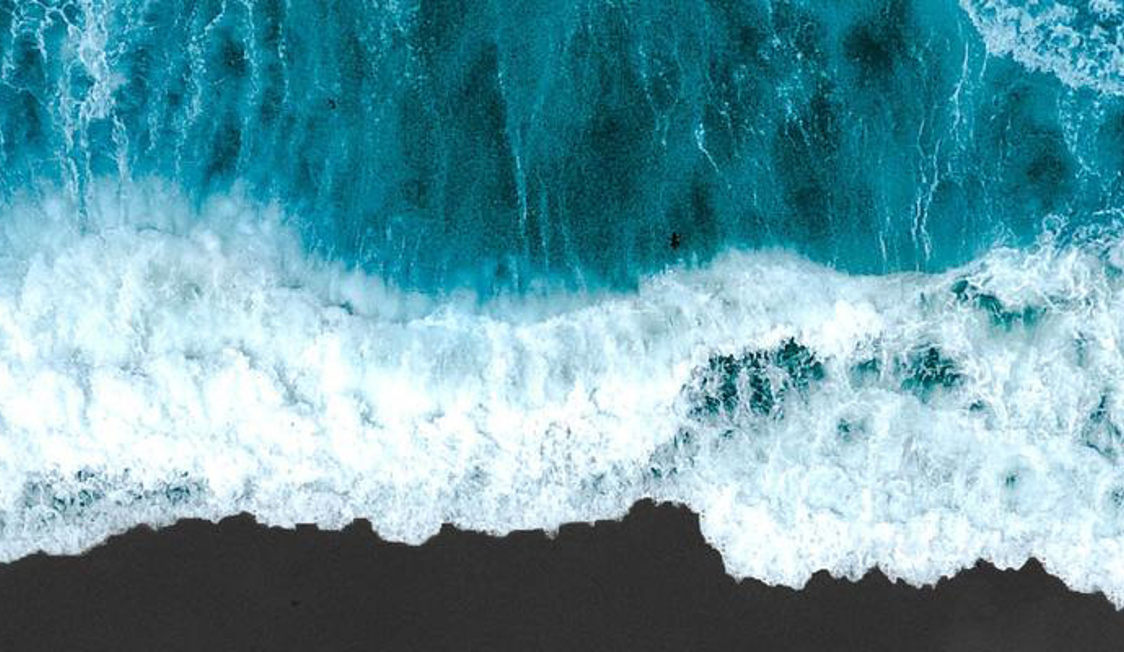 Böljor bryter mot kusten i fågelperspektiv, visar turkosfärgat hav omgivet av skummande vitt vatten.