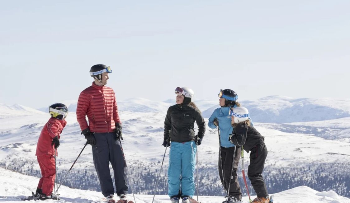 Familie på skiferie på snedækkede bjerge, iført farverige skijakker og hjelme, kigger ud over landskabet.
